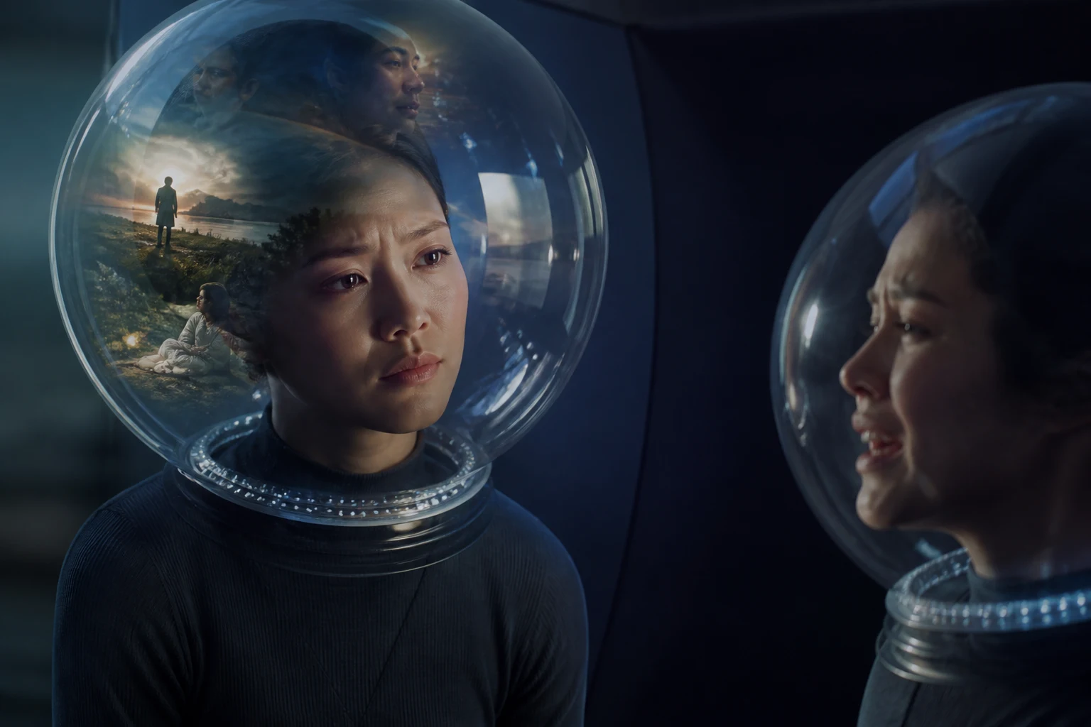 A listener watches a sorrowful speaker through layered reflections in two transparent helmets