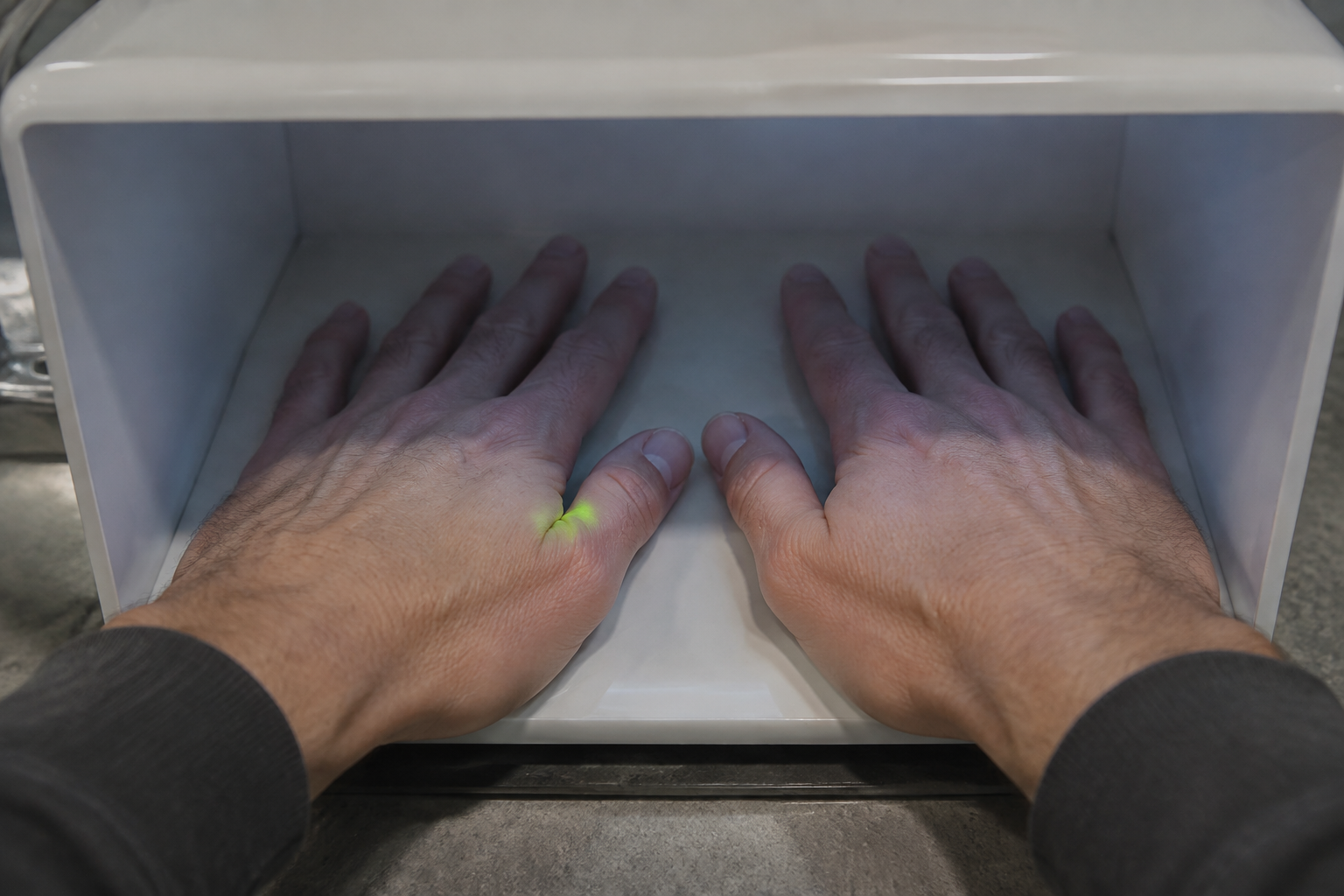 Close concept view of both hands inside a compact inspection hood, with a small yellow-green fluorescent patch near a thumb